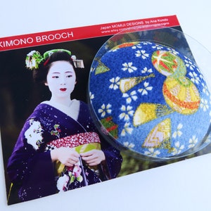 May include: A circular brooch featuring a blue fabric surface with a gold and white floral pattern, displayed on a card labeled KIMONO BROOCH. The card shows a photograph of a person in a dark purple kimono with traditional white makeup and hair ornaments.