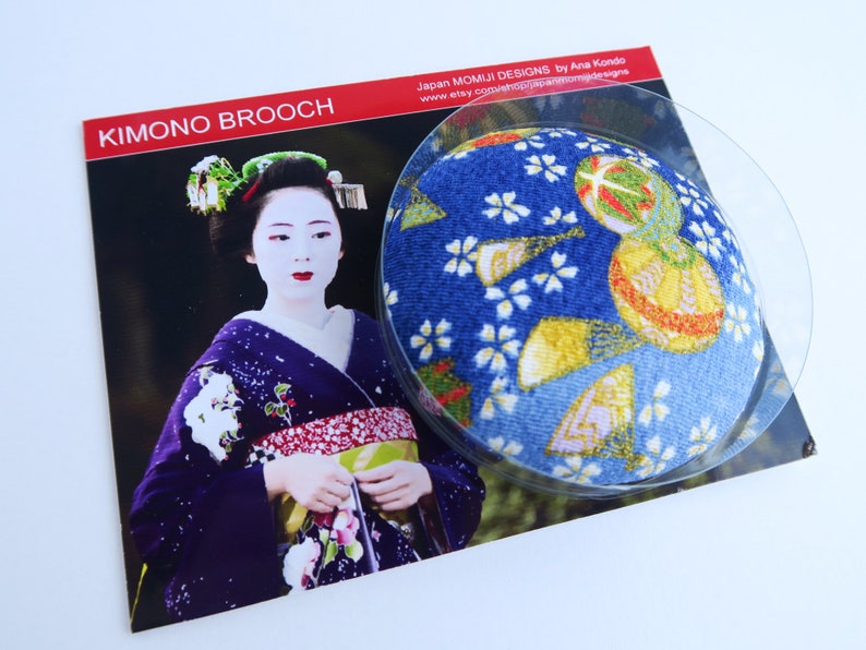 May include: A circular brooch featuring a blue fabric surface with a gold and white floral pattern, displayed on a card labeled KIMONO BROOCH. The card shows a photograph of a person in a dark purple kimono with traditional white makeup and hair ornaments.