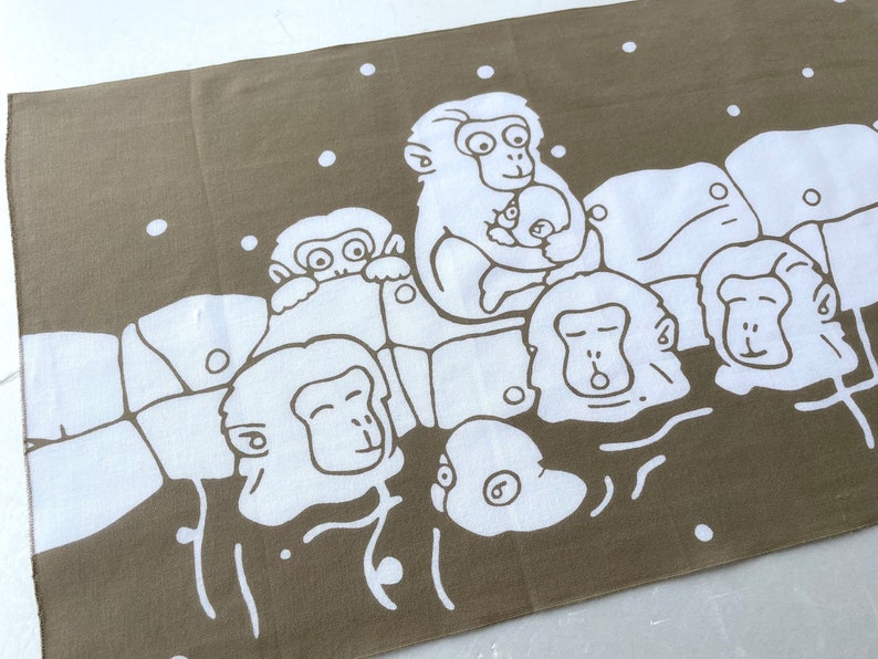 May include: Brown and white patterned towel featuring a group of cartoon monkeys relaxing in a hot spring. The monkeys are white with black outlines, and the background is brown. White dots represent falling snow.