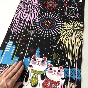 May include: Two white lucky cat figurines with black spots and pink accents are standing on a black background with a cityscape and fireworks. The cat on the left is wearing a green and white tie and the cat on the right is wearing a red and white polka dot scarf.