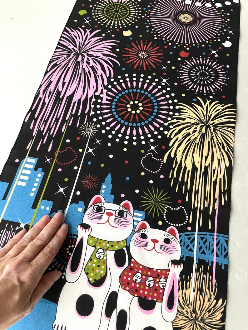 May include: Two white lucky cat figurines with black spots and pink accents are standing on a black background with a cityscape and fireworks. The cat on the left is wearing a green and white tie and the cat on the right is wearing a red and white polka dot scarf.