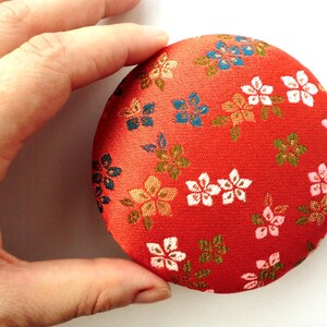 May include: A round, padded fabric pincushion featuring a vibrant red background with a repeating pattern of small, multi-colored floral blossoms in shades of white, pink, blue, and gold. The circular object has a soft, textured textile surface.