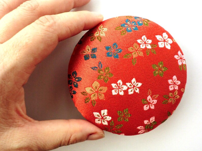 May include: A round, padded fabric pincushion featuring a vibrant red background with a repeating pattern of small, multi-colored floral blossoms in shades of white, pink, blue, and gold. The circular object has a soft, textured textile surface.