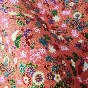 May include: A close-up of a pink fabric with a floral pattern. The fabric is covered in small flowers in shades of pink, red, blue, and white. The flowers are surrounded by green leaves and gold accents.