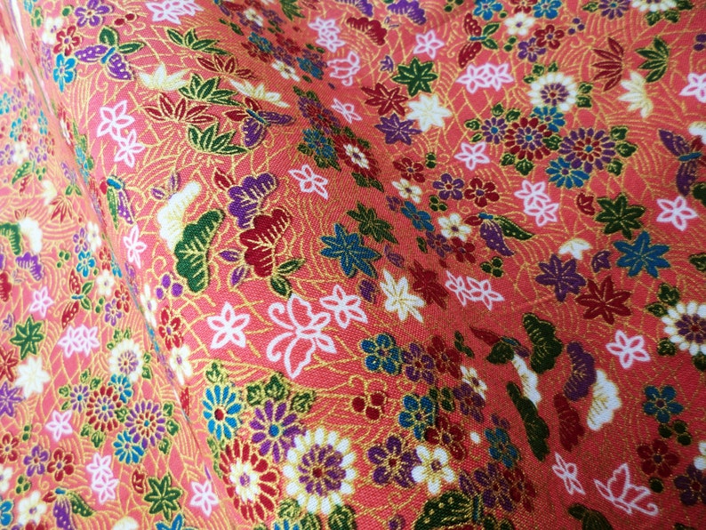May include: A close-up of a pink fabric with a floral pattern. The fabric is covered in small flowers in shades of pink, red, blue, and white. The flowers are surrounded by green leaves and gold accents.