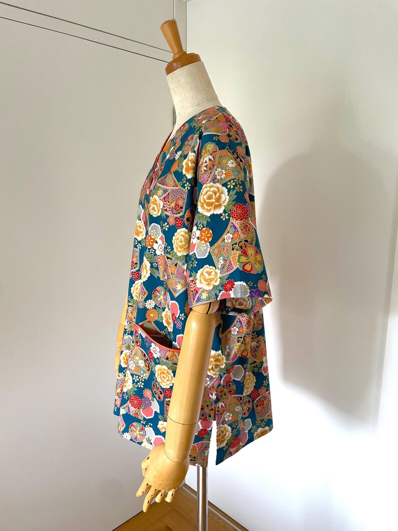 Puede incluir: Una camisa de manga corta azul turquesa con un estampado floral colorido. La camisa tiene cuello en V y dos bolsillos en la parte delantera.