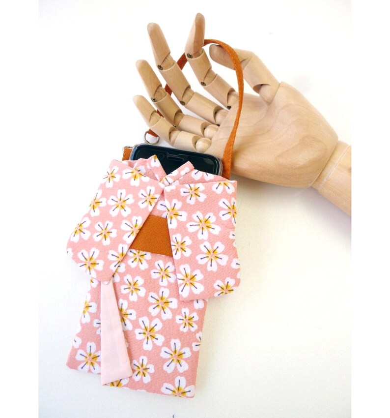 May include: A pink fabric phone pouch designed to resemble a kimono, featuring a white and yellow cherry blossom pattern. The pouch includes a solid orange obi-style band across the middle and a light pink fabric accent hanging from the side.