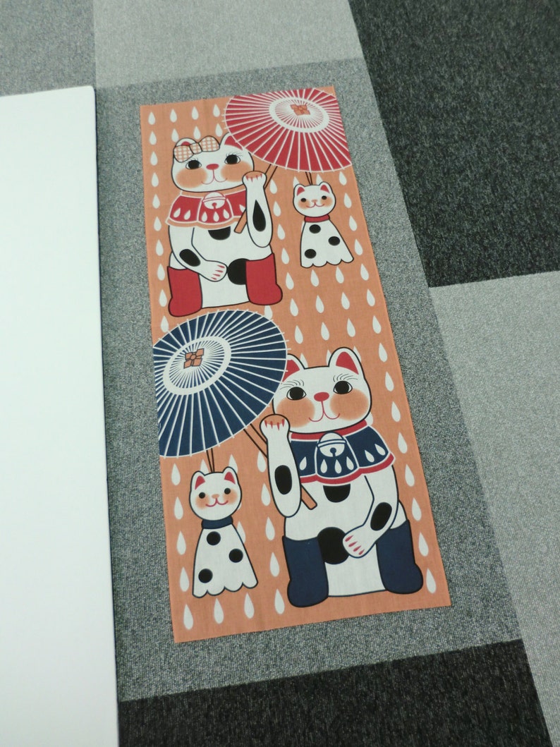 May include: A tan fabric panel with a repeating pattern of raindrops. Three white cats with black spots are holding umbrellas. The cats are wearing red, blue, and black clothing.