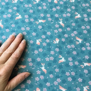 Peut inclure: Tissu bleu canard avec un motif r&eacute;p&eacute;t&eacute; de lapins blancs aux accents p&ecirc;che et de petites fleurs roses. Le tissu a un aspect textur&eacute;, adapt&eacute; &agrave; divers projets cr&eacute;atifs.