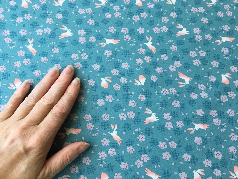 Peut inclure: Tissu bleu canard avec un motif r&eacute;p&eacute;t&eacute; de lapins blancs aux accents p&ecirc;che et de petites fleurs roses. Le tissu a un aspect textur&eacute;, adapt&eacute; &agrave; divers projets cr&eacute;atifs.