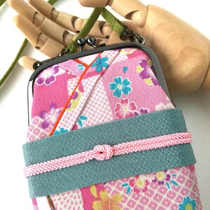 May include: A rectangular fabric pouch with a metal kiss-lock clasp, featuring a pink floral pattern with blue and yellow accents. A wide teal band wraps around the center, secured by a thin pink braided cord. It includes a green suede strap.