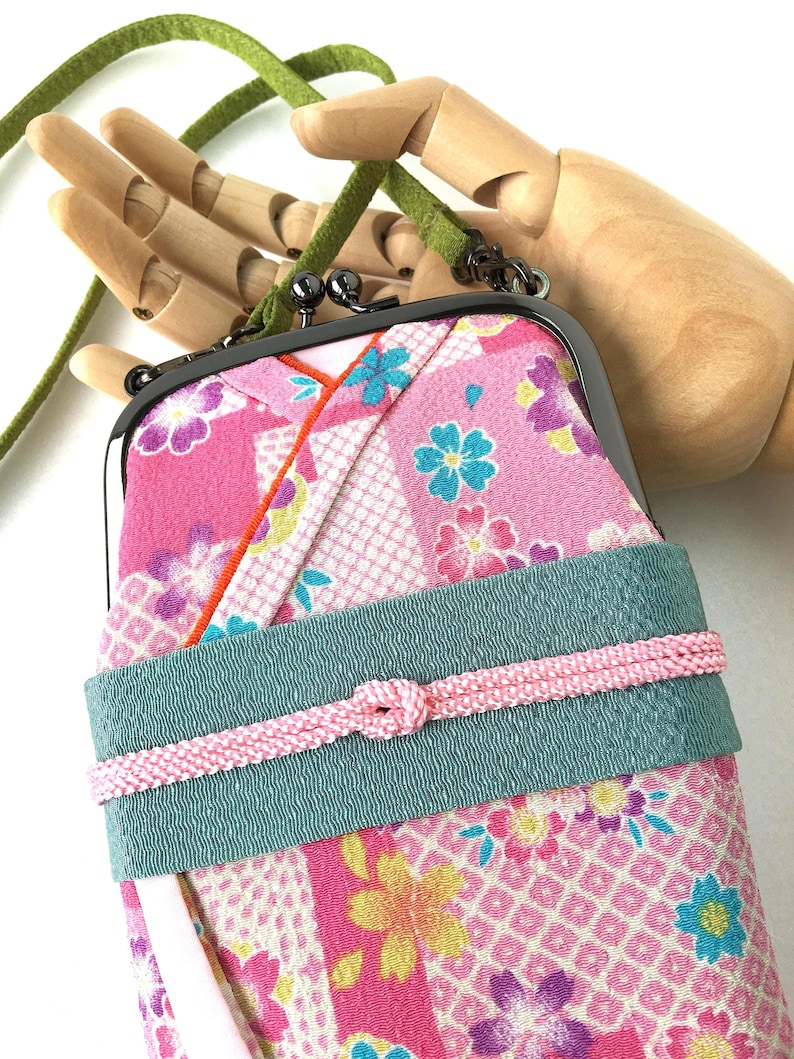 May include: A rectangular fabric pouch with a metal kiss-lock clasp, featuring a pink floral pattern with blue and yellow accents. A wide teal band wraps around the center, secured by a thin pink braided cord. It includes a green suede strap.