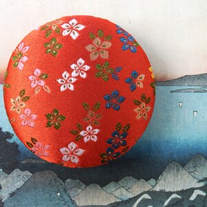 May include: A circular, padded object covered in vibrant red fabric featuring a repeating pattern of small, embroidered flowers in shades of white, pink, blue, and green. The surface has a textured, woven appearance with visible stitching details.