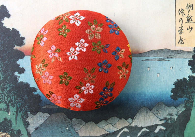 May include: A circular, padded object covered in vibrant red fabric featuring a repeating pattern of small, embroidered flowers in shades of white, pink, blue, and green. The surface has a textured, woven appearance with visible stitching details.