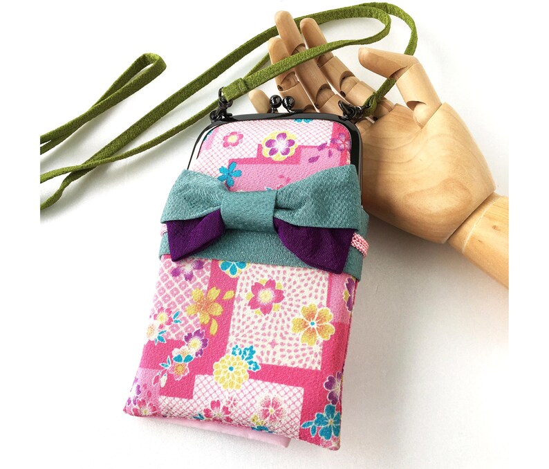 May include: A rectangular fabric pouch with a pink floral pattern featuring yellow and blue blossoms. The pouch has a black metal kiss-lock frame, a teal and purple fabric bow attached to the front, and a long green cord strap for carrying.