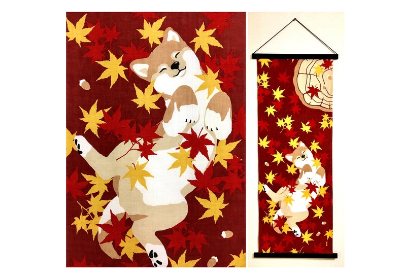 May include: A vertical fabric wall hanging featuring a tan and white Shiba Inu dog lying on its back amidst a scattering of red and yellow maple leaves on a deep red background. The design includes a tree stump graphic and a black hanging rod.