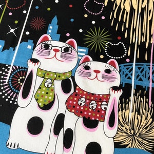 May include: Two white lucky cat figurines with black spots and pink accents, wearing a green and red scarf with bells, against a black background with fireworks and a cityscape.