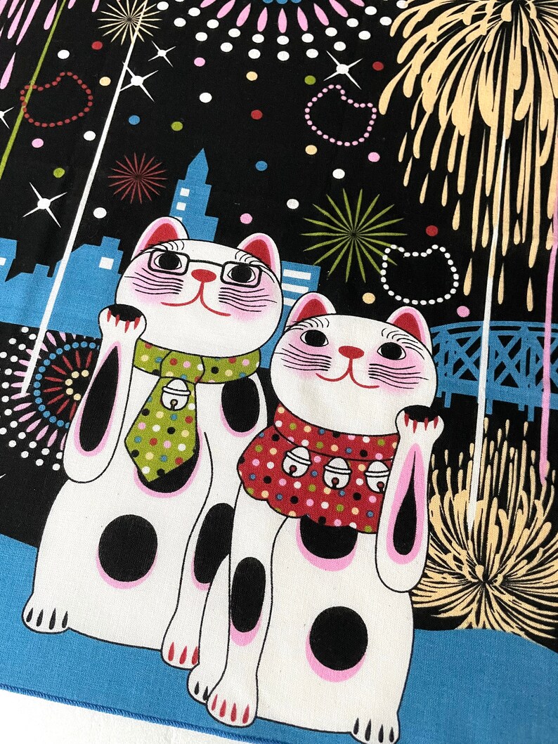 May include: Two white lucky cat figurines with black spots and pink accents, wearing a green and red scarf with bells, against a black background with fireworks and a cityscape.