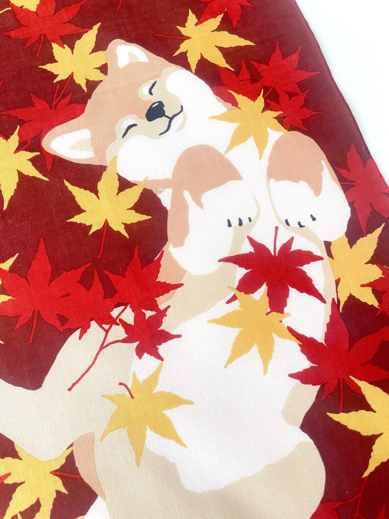 May include: A fabric textile featuring a tan and white Shiba Inu dog lying on its back with eyes closed, surrounded by a pattern of vibrant red and yellow maple leaves against a deep maroon background, creating a seasonal autumn-themed design.