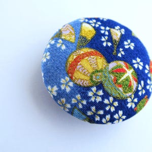 May include: A circular fabric-covered button featuring a vibrant blue background with a repeating pattern of small white cherry blossom flowers and larger, detailed golden and green decorative spheres with intricate line work and metallic accents.