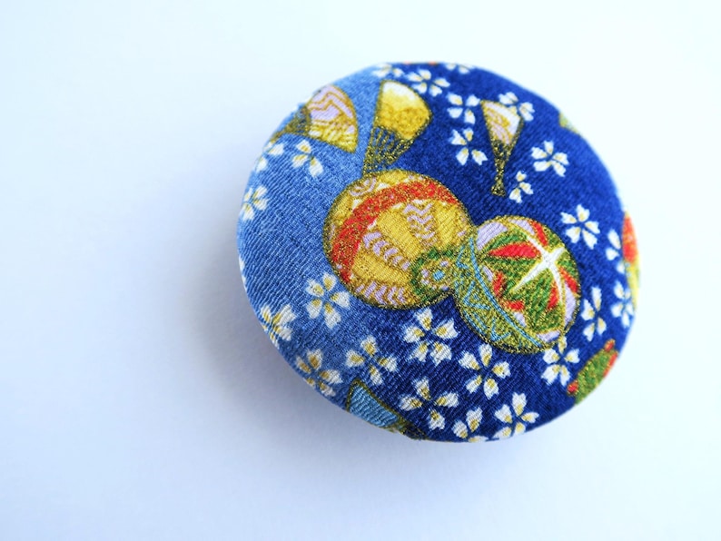 May include: A circular fabric-covered button featuring a vibrant blue background with a repeating pattern of small white cherry blossom flowers and larger, detailed golden and green decorative spheres with intricate line work and metallic accents.