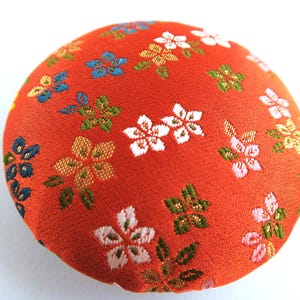 May include: A round, fabric-covered button featuring a vibrant orange background with a woven pattern of small, multi-colored flowers in shades of white, light pink, blue, and green. The surface has a textured, woven appearance throughout.