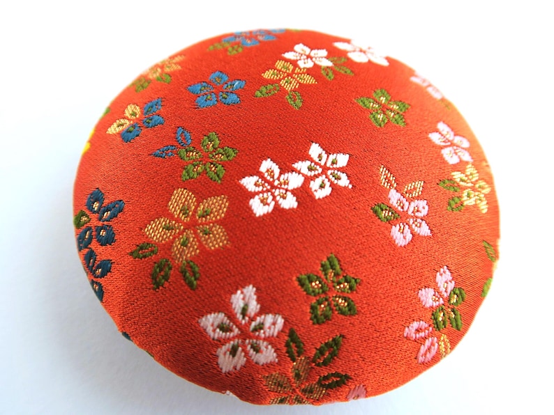 May include: A round, fabric-covered button featuring a vibrant orange background with a woven pattern of small, multi-colored flowers in shades of white, light pink, blue, and green. The surface has a textured, woven appearance throughout.