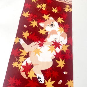May include: A rectangular fabric textile featuring a deep red background with a pattern of scattered yellow and red maple leaves. A light brown and white dog is depicted lying on its back among the leaves, with a tree stump graphic in the corner.