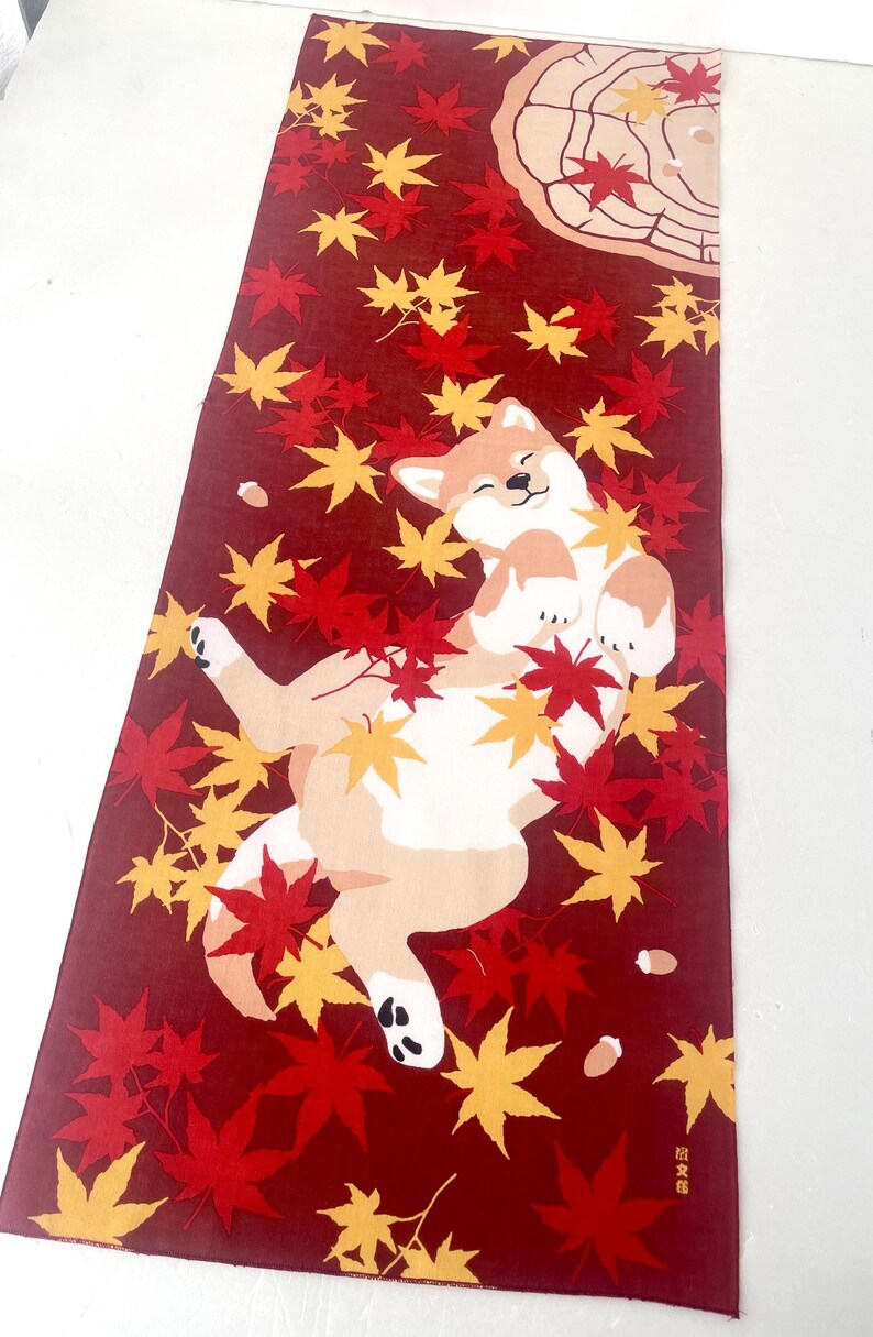 May include: A rectangular fabric textile featuring a deep red background with a pattern of scattered yellow and red maple leaves. A light brown and white dog is depicted lying on its back among the leaves, with a tree stump graphic in the corner.