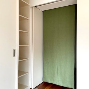 Puede incluir: Un armario blanco con puertas correderas y estantes. La puerta del armario est&aacute; abierta, revelando un divisor de habitaci&oacute;n de tela verde detr&aacute;s.