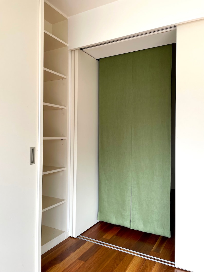 Puede incluir: Un armario blanco con puertas correderas y estantes. La puerta del armario est&aacute; abierta, revelando un divisor de habitaci&oacute;n de tela verde detr&aacute;s.