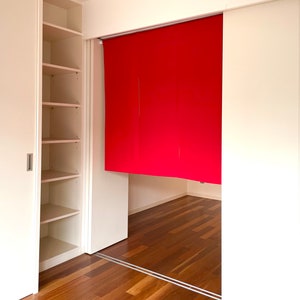 May include: A white closet with sliding doors, one door is open revealing empty shelves. The other door is closed and has a red fabric panel behind it. The floor is a light brown wood.