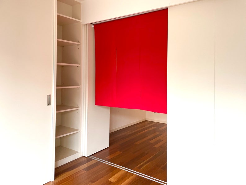 May include: A white closet with sliding doors, one door is open revealing empty shelves. The other door is closed and has a red fabric panel behind it. The floor is a light brown wood.