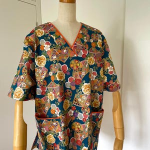 Puede incluir: Un top de uniforme m&eacute;dico azul turquesa con cuello en V y mangas cortas. La tela presenta un estampado floral y geom&eacute;trico colorido con detalles dorados. El top tiene un solo bolsillo en el lado izquierdo con un ribete naranja.