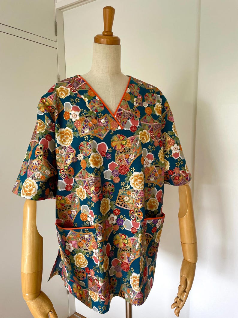 Puede incluir: Un top de uniforme m&eacute;dico azul turquesa con cuello en V y mangas cortas. La tela presenta un estampado floral y geom&eacute;trico colorido con detalles dorados. El top tiene un solo bolsillo en el lado izquierdo con un ribete naranja.