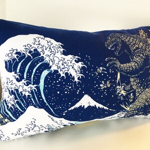 May include: A blue pillow with a white wave design and a Godzilla-like monster on the right side. The wave design is reminiscent of the Great Wave off Kanagawa, a famous Japanese woodblock print.