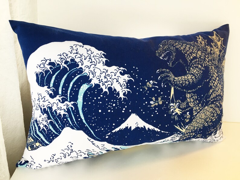 May include: A blue pillow with a white wave design and a Godzilla-like monster on the right side. The wave design is reminiscent of the Great Wave off Kanagawa, a famous Japanese woodblock print.