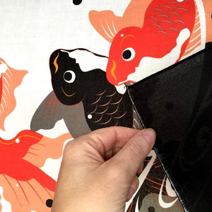 May include: A close-up of a hand holding a black fabric over a white fabric with a red and black fish design. The fish are stylized and have a traditional Japanese look.