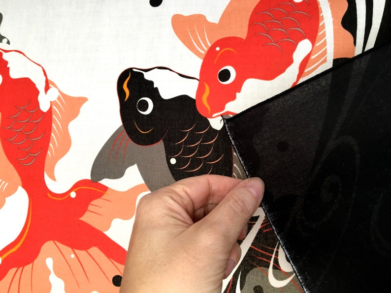 May include: A close-up of a hand holding a black fabric over a white fabric with a red and black fish design. The fish are stylized and have a traditional Japanese look.