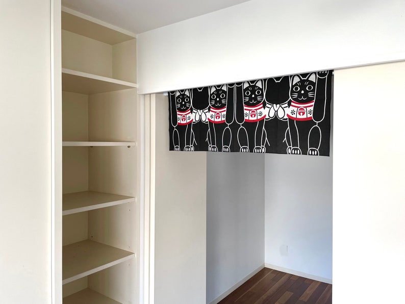 Puede incluir: Cortina japonesa Maneki Neko en blanco y negro en una puerta. La cortina presenta un patr&oacute;n repetido de gatos estilizados con las patas levantadas, con collares rojos. Los gatos est&aacute;n sobre un fondo negro.