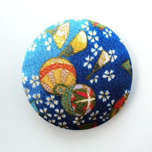 May include: A circular fabric-covered button featuring a deep blue background with a repeating pattern of small white cherry blossoms, golden fan shapes, and colorful spherical ornaments with red, green, and yellow geometric details.