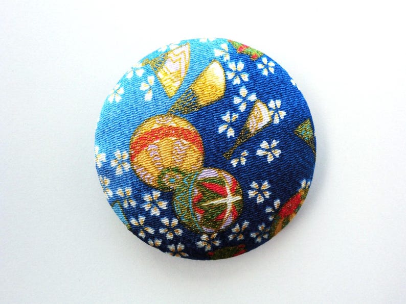 May include: A circular fabric-covered button featuring a deep blue background with a repeating pattern of small white cherry blossoms, golden fan shapes, and colorful spherical ornaments with red, green, and yellow geometric details.