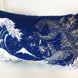 May include: A blue pillow with a white and blue print of Godzilla standing in front of a large wave and Mount Fuji.