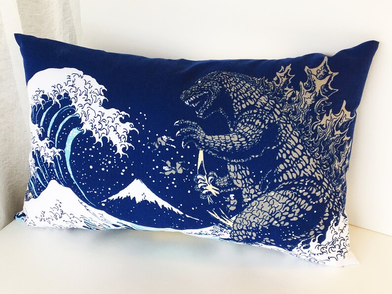 May include: A blue pillow with a white and blue print of Godzilla standing in front of a large wave and Mount Fuji.