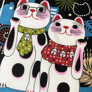 May include: Two white lucky cat figurines with pink cheeks and black eyes. The cats are wearing red and green scarves with white polka dots. The background is black with white polka dots and fireworks.