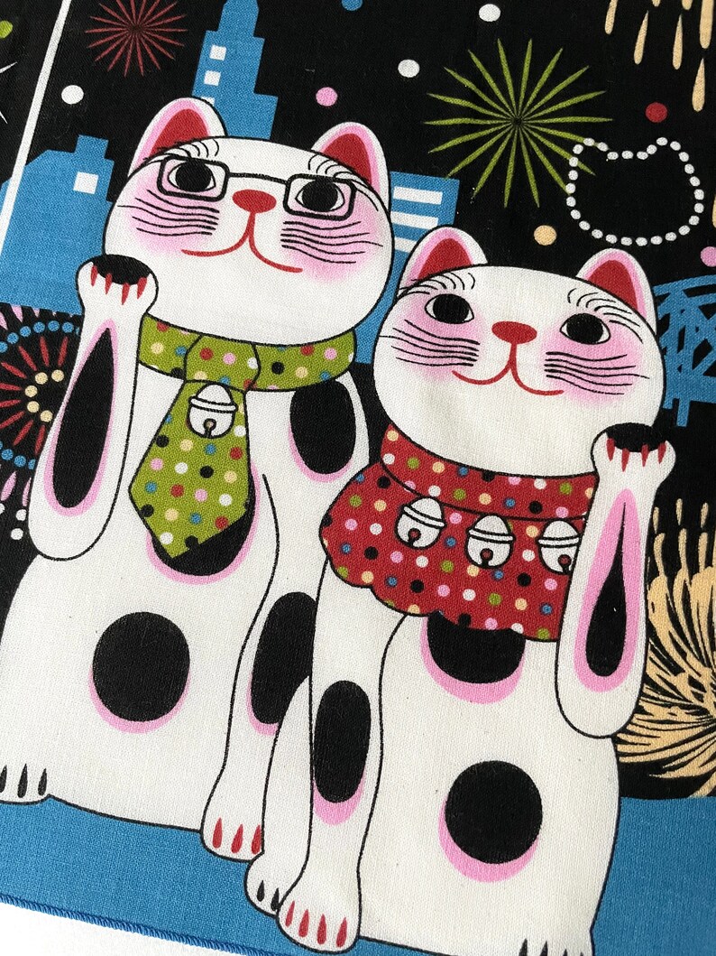 May include: Two white lucky cat figurines with pink cheeks and black eyes. The cats are wearing red and green scarves with white polka dots. The background is black with white polka dots and fireworks.