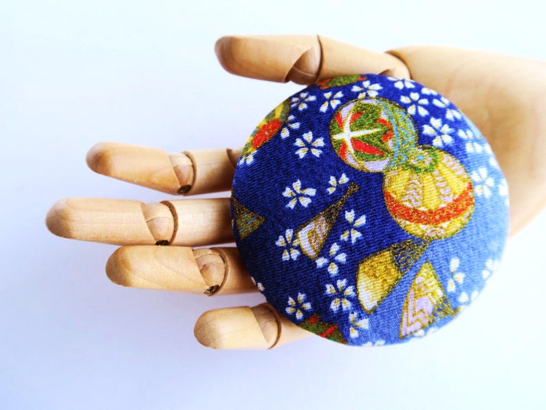 May include: A circular fabric-covered button featuring a deep blue background with a repeating pattern of white cherry blossoms, gold-accented folding fans, and spherical ornaments with red, green, and gold geometric details.