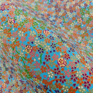 Puede incluir: Una tela de color azul claro con un patr&oacute;n denso y repetitivo de peque&ntilde;as flores de cerezo naranjas, rosas y moradas, intercaladas con motivos circulares dorados y tallos verdes finos, creando una superficie floral texturizada y colorida.