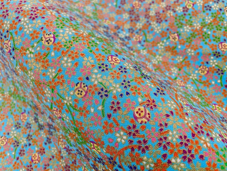Puede incluir: Una tela de color azul claro con un patr&oacute;n denso y repetitivo de peque&ntilde;as flores de cerezo naranjas, rosas y moradas, intercaladas con motivos circulares dorados y tallos verdes finos, creando una superficie floral texturizada y colorida.
