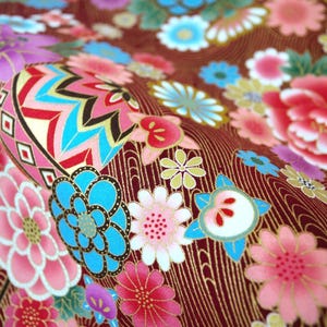 May include: A fabric featuring a deep red background with a repeating pattern of colorful flowers in shades of pink, blue, and white, interspersed with gold-toned wavy lines and a prominent geometric circular motif with blue and pink accents.